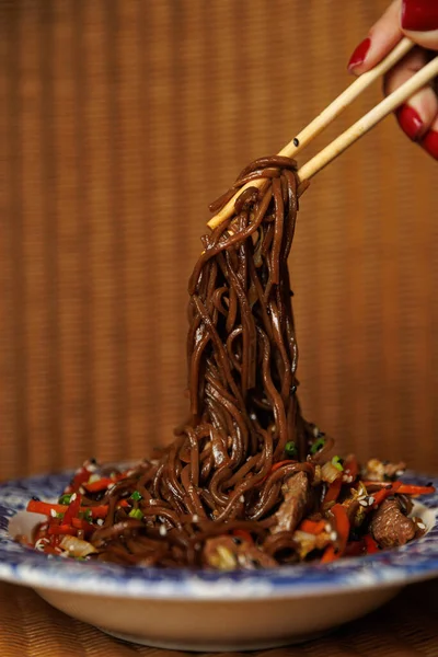 Bir el yemek çubuğu rengarenk sebzeler ve et parçalarıyla dolu kızarmış erişteleri kaldırıyor. Tabak, canlı renkler içeren dekoratif bir tabakta servis edilir..