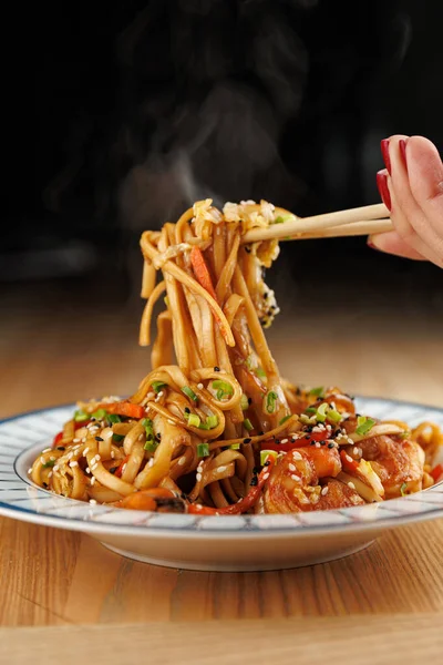 A hand holds chopsticks to grab a portion of steaming stir-fried noodles. The dish is filled with vegetables and shrimp, served on a white plate with a blue rim.