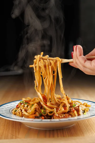 A hand holds chopsticks while lifting steaming noodles from a plate filled with colorful vegetables. The kitchen has a warm atmosphere, showcasing the joy of eating.