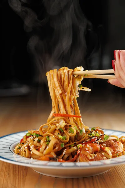 Steaming noodles topped with vegetables and sesame are lifted by chopsticks from a bright blue plate. The meal is set on a wooden table and captures a moment of enjoyment.