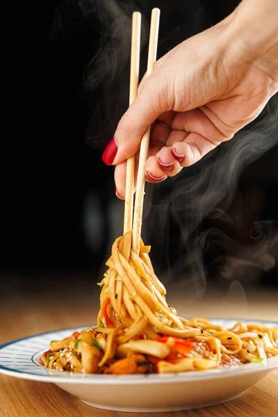 A hand holding chopsticks picks up steaming noodles from a bowl. Various vegetables and flavors add to the dish. This scene captures the enjoyment of Asian cuisine.