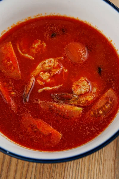 Bir kase kırmızı çorba deniz ürünleri ve sebzeler içerir, çeşitli tatlar içerir. Çorba zengin ve sağlıklı, tadını çıkarmaya hazır. Ayar, ahşap bir masanın üzerindeki tabağı vurguluyor..