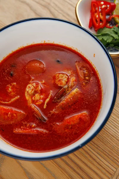 Bir kase zengin kırmızı deniz ürünleri çorbası ahşap bir masanın üzerine konur. Tabakta karides ve baharatlı et suyuyla çevrili diğer deniz ürünleri yer alıyor. Kırmızı biberli ve otlu bir tabak yemeği tamamlar..