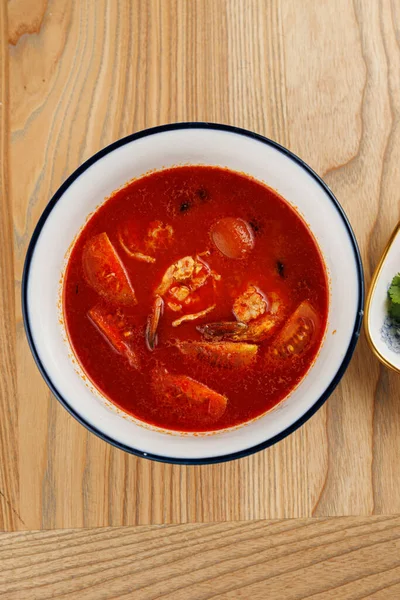 Karides ve sebzeli zengin kırmızı bir çorba ahşap bir masada bir kaseye konur. Çorbada çeşitli tatlar ve parlak renkler var..
