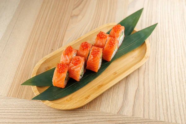 Balık yumurtası ile kaplı dilimlenmiş somon balığı ruloları bambu tabağına yerleştirilmiş yeşil yapraklar üzerinde güzelce düzenlenir. Ayar tabağı tamamlayan hafif ahşap bir masa içeriyor..