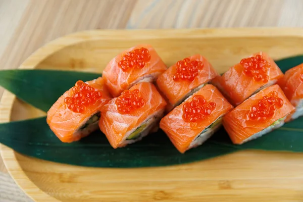 Taze hazırlanmış suşi ruloları ahşap bir tabakta sunulur. Her rulo somon dilimleri içeriyor ve üzerinde parlak kırmızı balık yumurtası var. Yeşil yapraklar renk ekler.