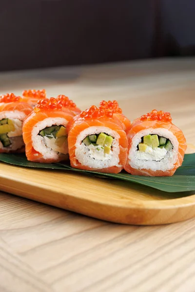 Somon, salatalık ve avokado ile dolu taze suşi ruloları ahşap servis tabağında oturur. Parlak turuncu somon ve malzemeler yemeğe renk katar..