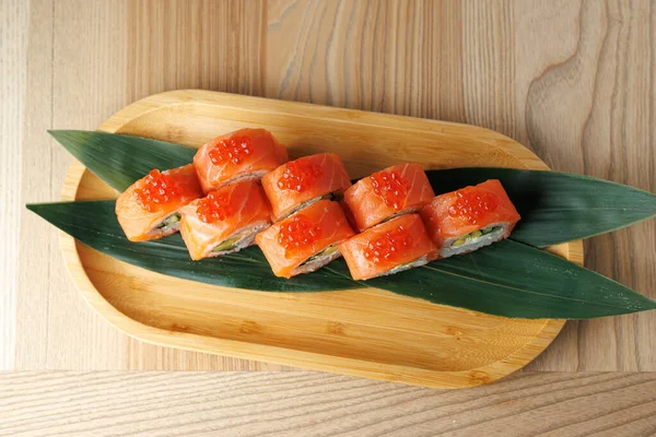 Bir tabakta somon dilimleri ve kırmızı havyarla kaplı suşi ruloları var. Rahat bir yemek atmosferinde bambu yaprakları üzerinde sunuluyor..