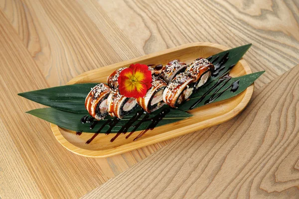 Suşi ruloları yılan balığı ile dolu ve susam yeşil yapraklar üzerinde ahşap bir tabakta dinleniyor. Parlak bir çiçek Japon mutfağına odaklanmış bir restoranda yemeği süslüyor..