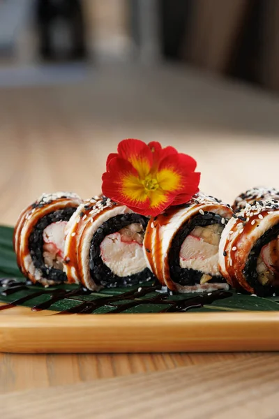 Deniz ürünleriyle dolu suşi ruloları bambu tabakta düzgünce düzenlenir. Canlı bir çiçek sunuma dekoratif bir dokunuş ekler. Dekor, çağdaş bir yemek havası veriyor..
