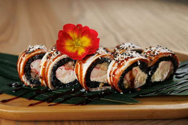 Taze suşi ruloları, susam tohumları ve serpiştirilmiş sosla ahşap bir tabakta güzelce düzenlenir. Renkli bir çiçek sunuma renk katar.
