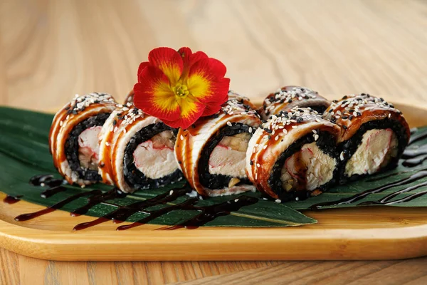 Suşi ruloları ahşap bir tabakta güzelce düzenlenir. Ekmeklerde çeşitli dolgular var ve üzerinde susam tohumları ve parlak bir çiçek var. Bir tutam sos tat katar..