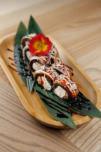 Suşi ruloları yeşil yapraklarla kaplanmış, çiçeklerle süslenmiş ve sosla süslenmiş ahşap bir tabakta düzenlenir. Sahne rahat bir restoranda geçiyor..