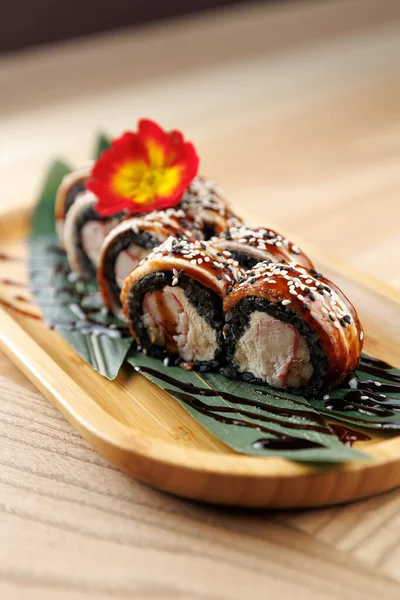 Suşi ruloları bambu tabakta güzelce düzenlenir. Tabakta siyah pirinç, deniz ürünleri ve dekorasyon için bir çiçek garnitürü yer alıyor. Sıradan bir yemek ortamında servis edilmiş..