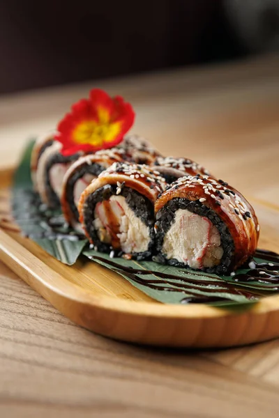 Suşi ruloları ahşap bir tabağa düzgünce yerleştirilir. Tabakta çeşitli malzemeler yer alıyor ve bir çiçekle süsleniyor. Dekorlar rahat bir restoran atmosferi sergiliyor..
