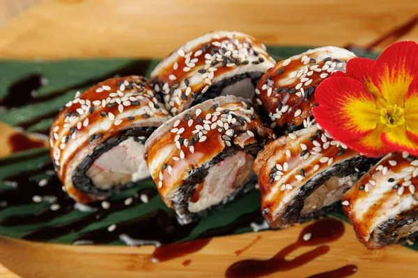 Suşi ruloları üzerine susam tohumları serpiştirilmiş ahşap bir tepside düzenlenir. Bir çiçek ekrana biraz renk katar. Bu yemek tatları ve sunumu vurgular.