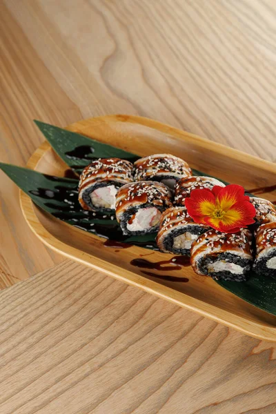 Suşi yuvarlakları yeşil yapraklı ahşap bir tepside ve çiçek süslemeli. Bir sos serpintisi bu Japon yemeğinin işçiliğini vurgularken sunumu güçlendirir..