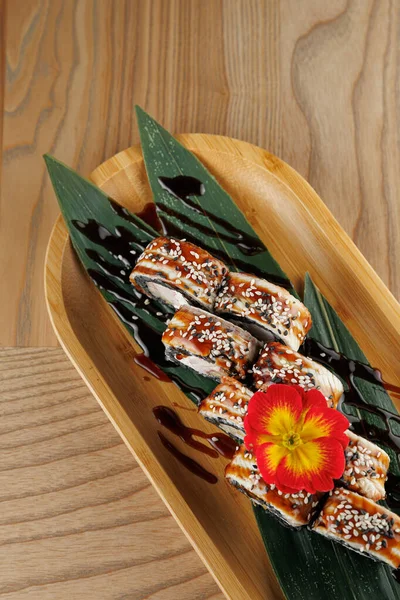 Bir bambu tepsisinde soslu ve susamlı pirinç ve yosun içeren suşi ruloları vardır. Bir çiçek renk katar, sunumu ahşap bir masada güçlendirir..