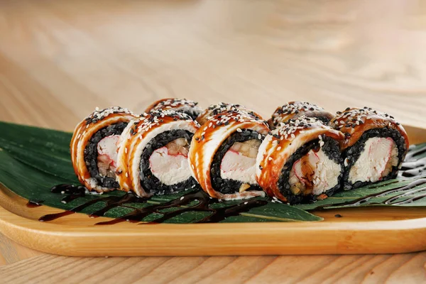 Sushi rolls are arranged neatly on a wooden plate, featuring rice, seaweed, and various fillings. Green leaves add a touch of freshness. A dark sauce is drizzled over the top.