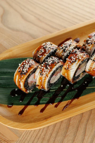 Üzerinde yılan balığı ve susam tohumları olan suşi ruloları ahşap bir tabakta bir yaprağın üzerinde duruyor. Kurulumda ahşap bir masa yer alıyor ve tabağın sunumunu vurgulayan bir sos serpintisi de yer alıyor.