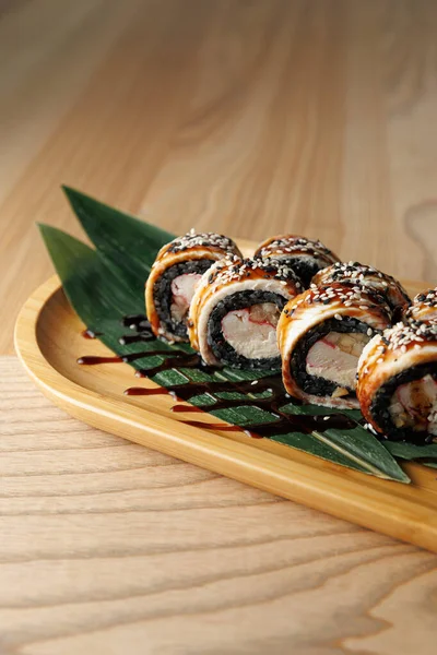 Suşi ruloları ahşap bir tabakta yeşil yapraklarla ve üzerine sos serpiştirilmiş bir şekilde düzgünce düzenlenir. Bu modern yemek sahnesinde Japon mutfağından popüler bir yemek sergileniyor..