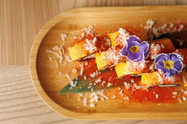 Tahta bir tabakta çeşitli suşi dürümleri. Ekmekler yenilebilir çiçeklerle kaplı ve yengeç eti serpiştirilmiş. Çanak canlı bir sunum sergiliyor.