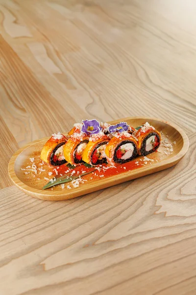 Tahta tabakta düzgünce dizilmiş birkaç suşi rulosu var. Ekmekler parlak renkler ve süslemeler içeriyor, onları görsel olarak çekici kılıyor. Ayarlar yemeğin sanatsallığını vurgular.