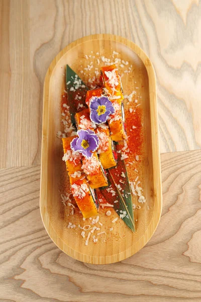 Suşi ruloları renkli malzemelerle kaplı ve yenilebilir çiçeklerle süslenmiş bir bambu tabakta sunulur. Dokuların ve tatların karışımı çeşitli mutfak geleneklerini temsil eder..