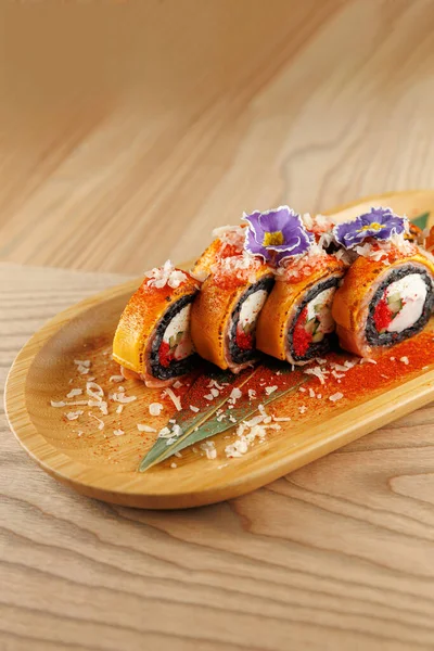 Suşi ruloları ahşap bir tabakta sergilenir. İçeriğinde canlı çiçekler ve malzemelerin karışımı var. Ayar, yemeğin dikkatli sunumu ve hazırlanmasını vurgular.