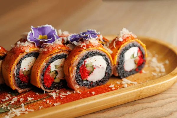 Taze suşi ruloları ahşap bir tabakta güzelce düzenlenir. Tabakta, üzerinde balık, krem peynir ve yenilebilir çiçekler bulunan canlı renkler gündelik bir şekilde servis ediliyor..