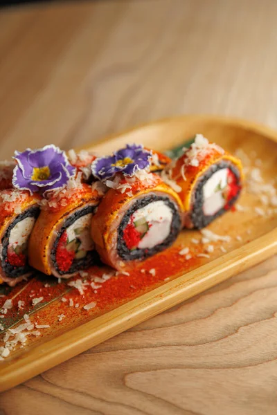 Suşi ruloları, yenilebilir çiçeklerle kaplı ve pullarla süslenmiş ahşap bir tabakta sergilenir. Sahne, Japon mutfağının bir yemek alanındaki sanatını yansıtıyor..