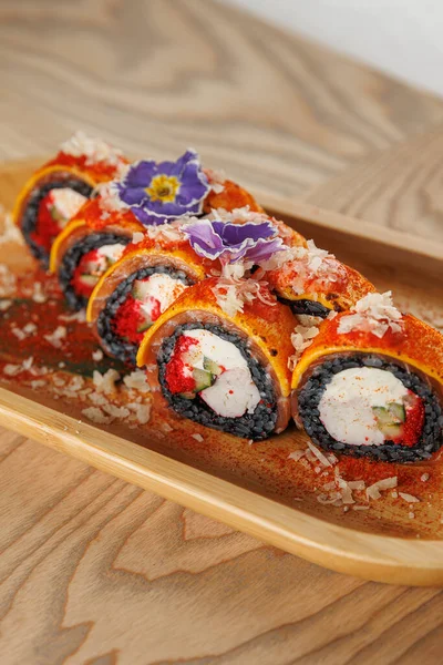 Suşi ruloları ahşap bir tabakta güzelce düzenlenir. Ekmekler renkli malzemeler içeriyor ve üzeri pullar ve çiçeklerle kaplı. Arka planda bir yemek alanı görülebilir..
