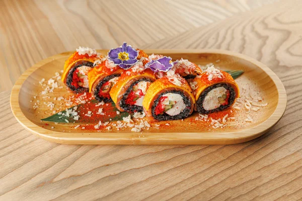 Suşi ruloları ahşap bir tabakta güzelce düzenlenir. Tabakta canlı malzemeler, taze sebzeler ve çekici bir sunum için eşsiz süsler yer alıyor..