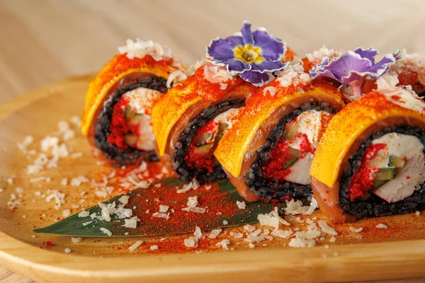 Suşi ruloları ahşap bir tabakta güzelce düzenlenir. Ekmekler sarı malzemelerle kaplı ve mor çiçeklerle süslenmiş. Tabakta baharat ve garnitür var..