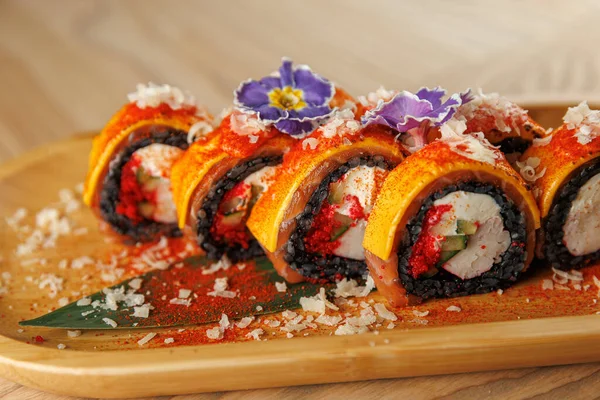 Ahşap bir tabağa yerleştirilmiş renkli suşi ruloları baharat ve yenilebilir çiçeklerle kaplı malzemelerin katmanlarını gösteriyor. Bu yemek Japon mutfak sanatını sergiliyor..