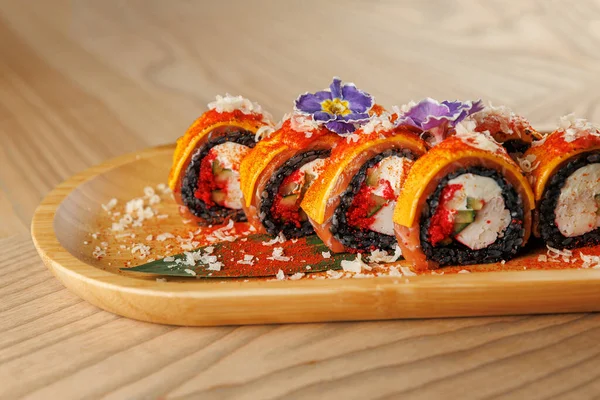 Farklı dolgularla suşi ruloları ahşap bir tabakta düzgünce düzenlenir. Parlak malzemeler ve süslemeler renk katar. Akşam yemeği ya da öğle yemeği saatinde bir restoranda olması muhtemel..
