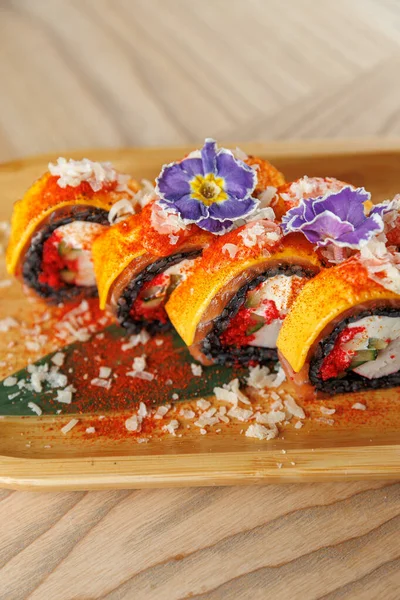 Renkli suşi ruloları ahşap bir tabakta güzelce düzenlenir. Ekmeğe yenilebilir çiçek ve baharat katılmış deniz ürünleri ve sebzeler katılıyor..