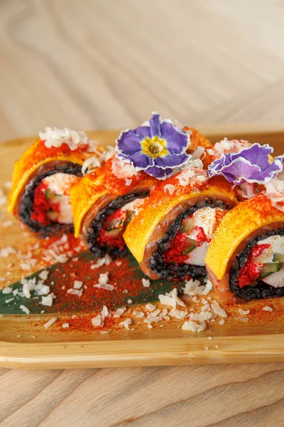 Renkli suşi ruloları ahşap bir tabakta güzelce düzenlenir. Her rulo sebze, çiçek ve çeşni ile kaplıdır, aroma ve sunum karışımı sunar..