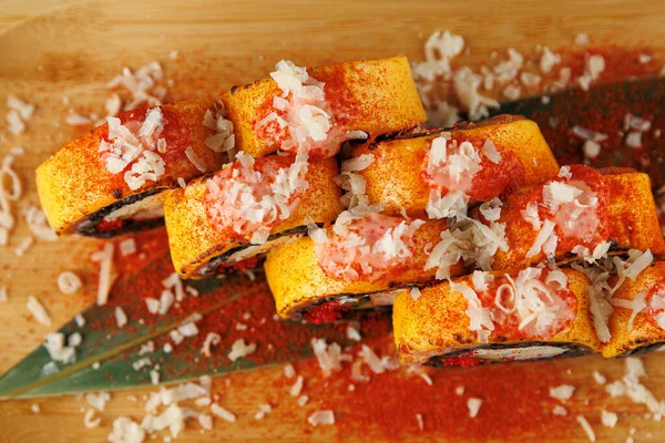 Suşi ruloları ahşap bir yüzeye istiflenmiş. Üzeri baharat ve parçalanmış malzemelerle kaplı, canlı renkler ve dokular sergileniyor. Sunuma dekoratif bir yaprak ekler.