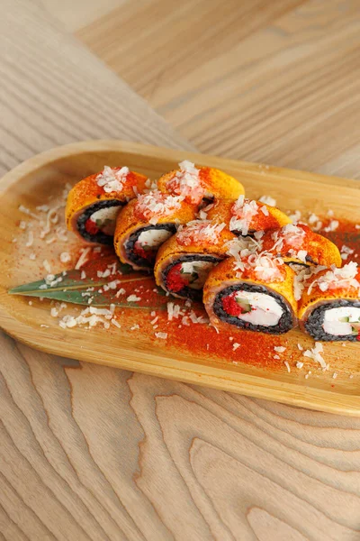 Renkli malzemelerle suşi ruloları ahşap bir tabakta oturur. Süsler ve soslar sunumu güçlendirir. Bu yemek, parlak bir ortamda sergileniyor, mutfak sanatı sergileniyor.