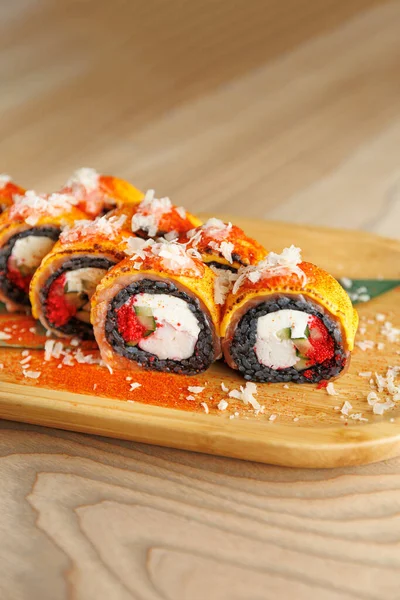 Ahşap bir tahtanın üzerinde, canlı suşi, kaliteli bir mutfakta hazırlanmış krem peynir ve sebzeler de dahil olmak üzere çeşitli malzemeler sergiliyor. Farklı renkler heyecan getirir.