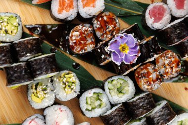 Tahta bir tahtanın üzerinde çeşitli suşi dürümleri var. Ekmekler arasında avokado, salatalık, ton balığı ve susam tohumlarıyla süslenmiş somon balığı ve bir çiçek var..
