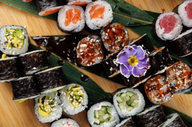 Suşi ruloları ahşap bir yüzeyde düzgün bir şekilde dizilir, somon ve avokado gibi farklı malzemeler sergilenir. Küçük çiçekler yemeğe dekoratif bir dokunuş ekler..