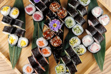 Suşi ruloları yeşil yapraklı ahşap bir tepside düzenlenir. Tabakta taze balık ve sebzelerin vurgulandığı çeşitli suşi türleri yer alıyor. Her rulo farklı dolgu ve malzemeler içeriyor..