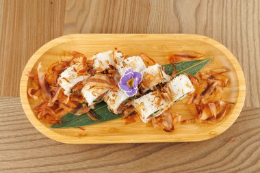 Ahşap bir tabakta suşi ruloları, bonito taneleri ve küçük bir çiçek bulunur. Dekor parlak, taze malzemelerin altını çiziyor ve yemeğin dikkatlice sunulmasını sağlıyor..