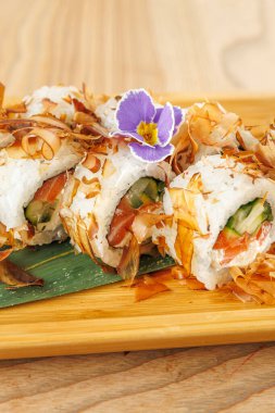 Taze sebzelerle dolu suşi ruloları ahşap bir tabakta yatıyor. Yoğun bir restoranın parlak ortamında bir çiçek yemeğe renk katar..