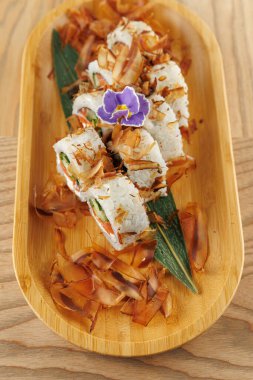 Suşi ruloları bambu tabağa konur. Kepekli malzemeler ve mor yenilebilir bir çiçek sunuma detay ekler. Bu sahne Japon mutfak sanatının bir karışımını gösteriyor..