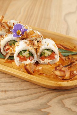 Tahta bir tabakta taze sebze ve balıktan yapılmış suşi ruloları. Çiçek ve çıtır çıtır gevreklerle süslenmiş bu yemek, dokuların ve tatların karışımını ön plana çıkarıyor..