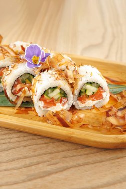 Taze suşi ruloları ahşap bir tabakta güzelce düzenlenir. Ekmeğin içinde salatalık ve somon var. Üstünde bonito gevreği ve çiçekle süslenmiş..