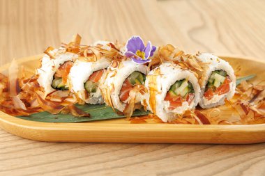 Salatalık ve somon dolu suşi ruloları ahşap bir tabakta düzenlenir. Tabakta bonito pulları ve küçük bir çiçek var. Japon mutfağını bir restoran ortamında sergiliyorlar..